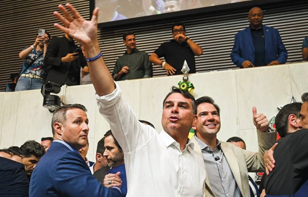 Jair Bolsonaro, de camisa branca, acena com a mão direita levantada, em meio a uma multidão. Ao seu lado, um homem de blazer bege sorri e faz um sinal de positivo. Outros homens observam de um balcão superior, incluindo um de blazer azul. O ambiente parece ser um evento político ou público, com pessoas filmando e fotografando.
